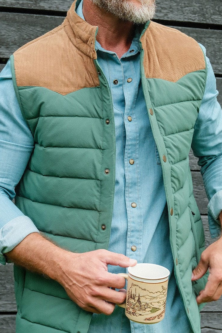 Man wearing a green puffer vest over a blue shirt, holding a paper cup against a wooden wall.